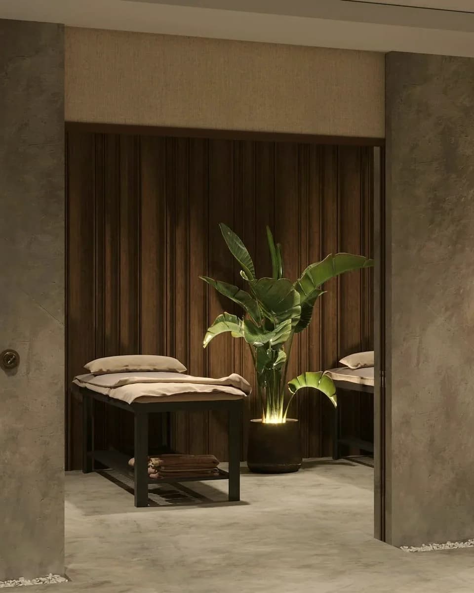 A clinic treatment room seen through a doorway, with a made bed, folded towels and a lit plant against dark timber.